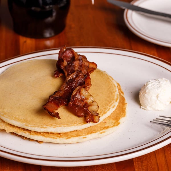 Two pancakes topped with crispy bacon and a scoop of butter on a white plate.