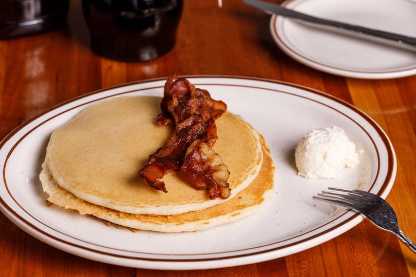 Two pancakes topped with crispy bacon and a scoop of butter on a white plate.