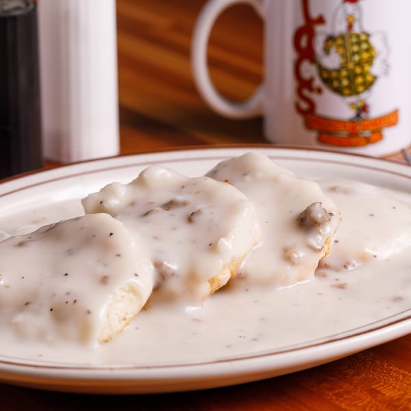 Three biscuits covered in creamy sausage gravy on a white plate, with a coffee mug in the background.