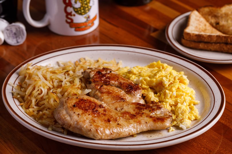 A breakfast plate with grilled chicken, scrambled eggs, hash browns, and toast on a wooden table.