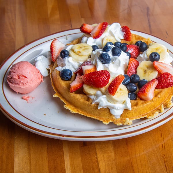 A waffle topped with whipped cream, strawberries, blueberries, and bananas, with a scoop of pink ice cream.