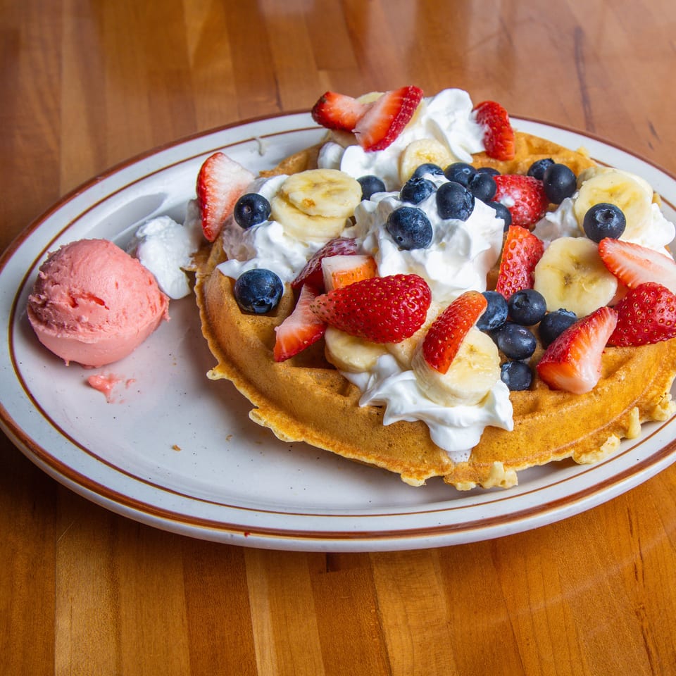 A waffle topped with whipped cream, strawberries, blueberries, and bananas, with a scoop of pink ice cream.