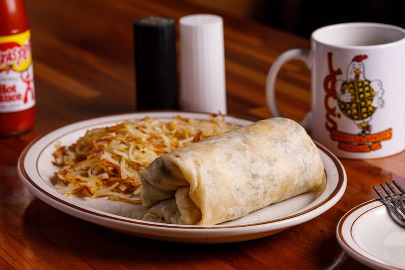 Breakfast burrito and hash browns on a plate with hot sauce, salt, pepper, and coffee.
