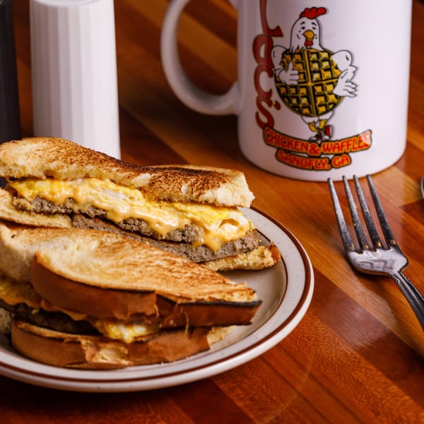 Breakfast sandwich with egg, cheese, and sausage on toast, with a branded mug, fork, and knife.