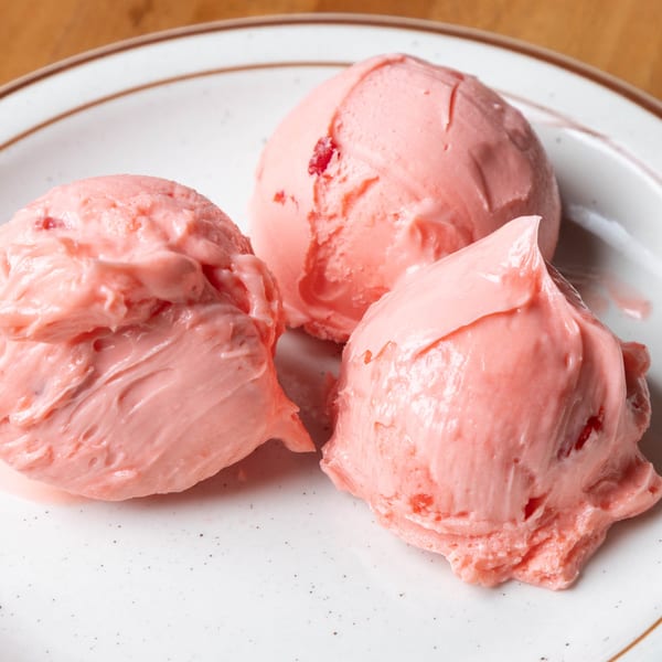 Three scoops of pink strawberry ice cream with fruit chunks on a white plate with a brown rim.