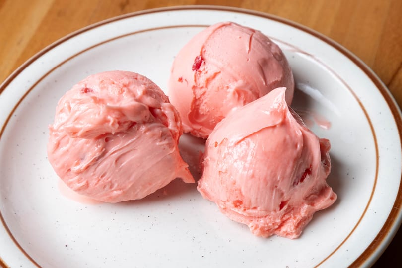 Three scoops of pink strawberry ice cream with fruit chunks on a white plate with a brown rim.