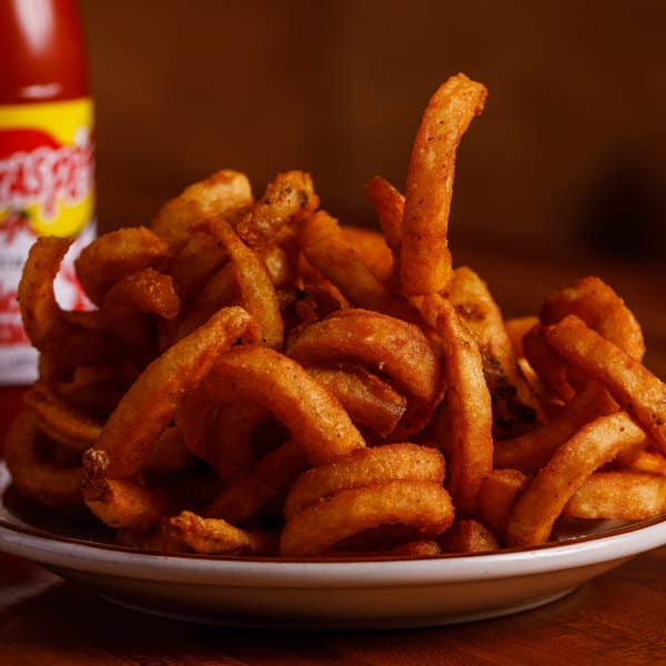 A plate of golden curly fries with a bottle of Texas Pete hot sauce in the background.