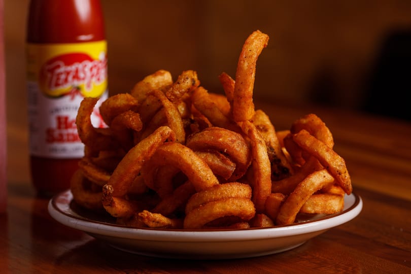 A plate of golden curly fries with a bottle of Texas Pete hot sauce in the background.