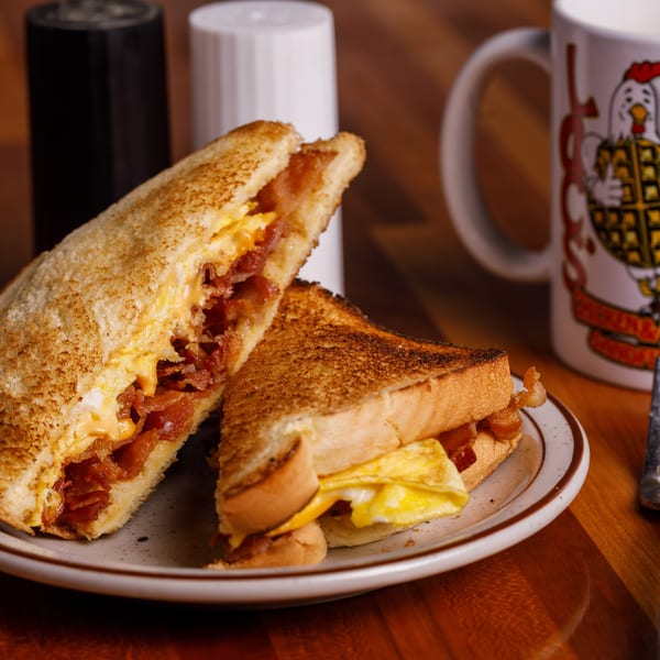 A breakfast sandwich with bacon, egg, and cheese, cut in half, on a plate with a coffee mug.