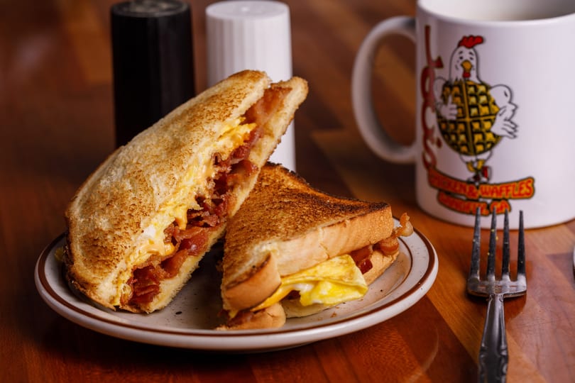 A breakfast sandwich with bacon, egg, and cheese, cut in half, on a plate with a coffee mug.