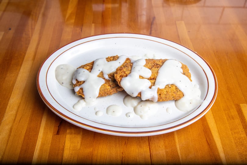 Two crispy fried chicken cutlets covered in creamy white gravy on a white plate.