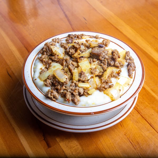 A bowl of creamy grits topped with browned sausage crumbles and caramelized onions on a wooden table.