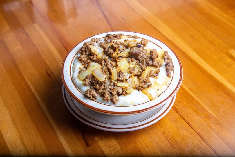 A bowl of creamy grits topped with browned sausage crumbles and caramelized onions on a wooden table.