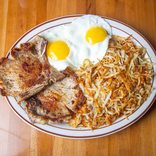 Two grilled pork chops, two fried eggs, and a pile of hash browns on a white plate.