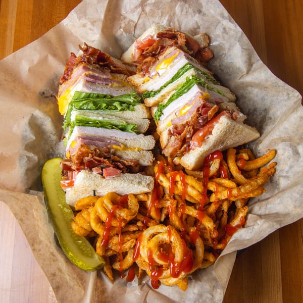 Club sandwich with bacon, lettuce, tomato, and egg, served with curly fries and a pickle.