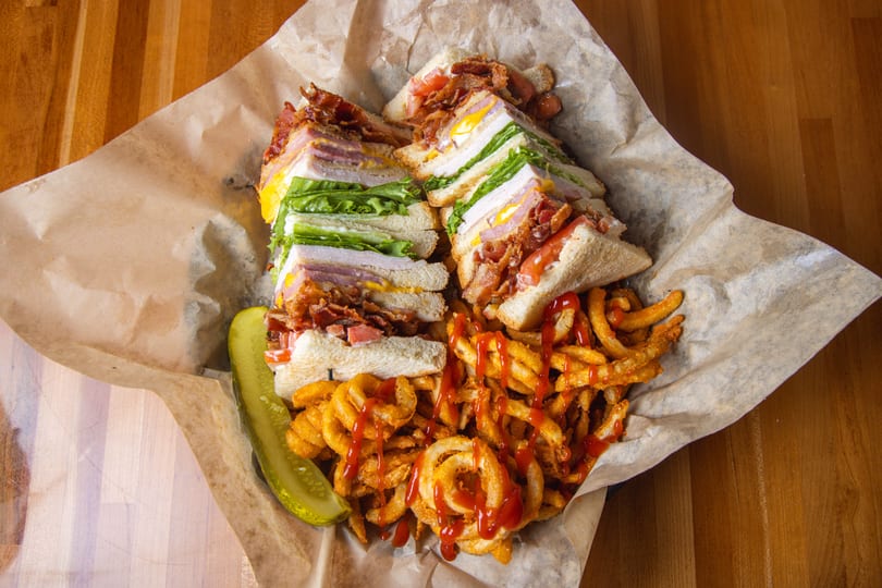 Club sandwich with bacon, lettuce, tomato, and egg, served with curly fries and a pickle.