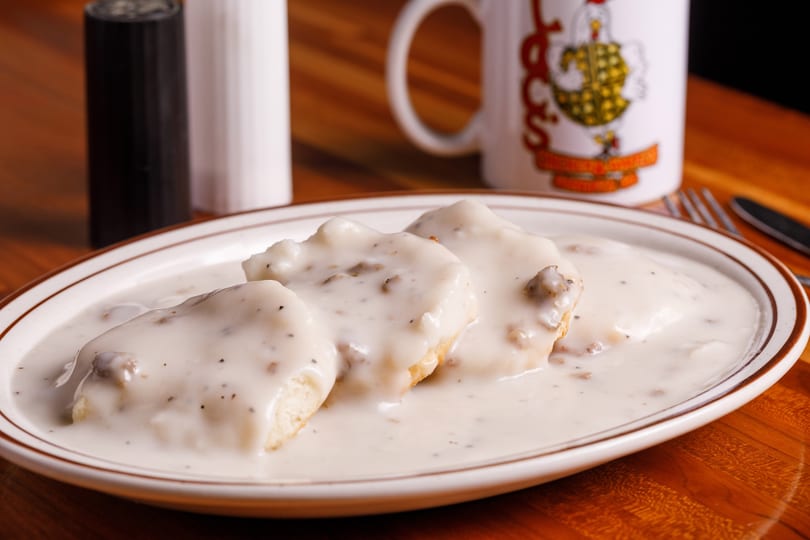 Three biscuits covered in creamy sausage gravy on a white plate, with a coffee mug in the background.