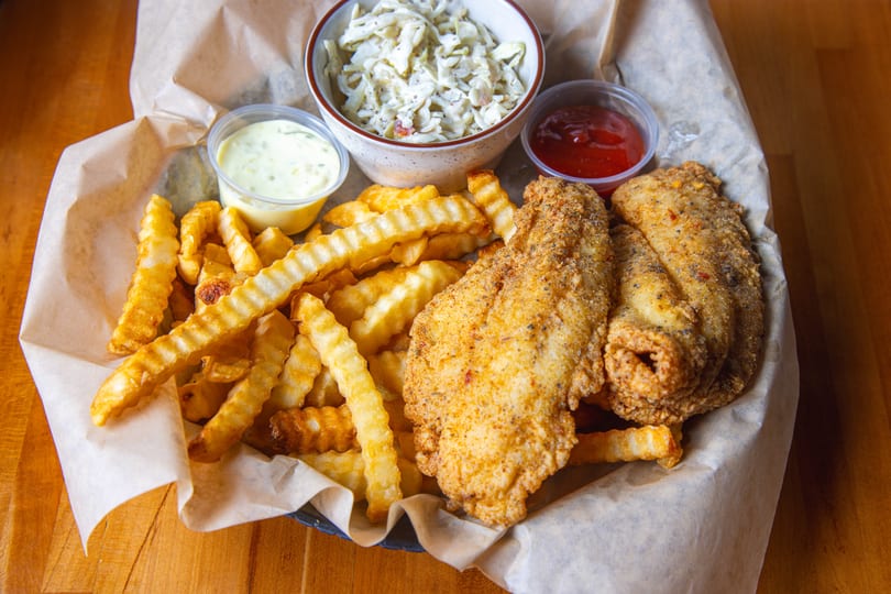 Fried fish and crinkle-cut fries with coleslaw, tartar sauce, and ketchup in a basket.