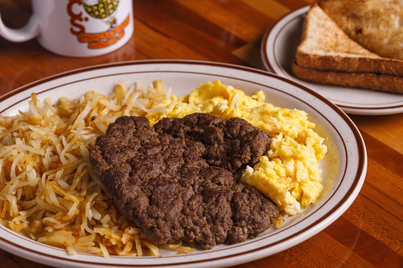 A breakfast plate with scrambled eggs, hash browns, and sausage patties, with toast and a mug in the background.