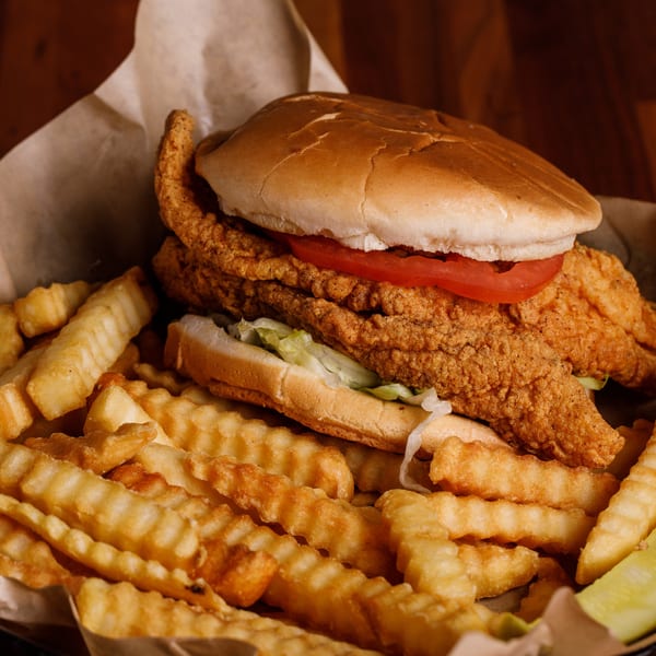 Crispy fried fish sandwich with lettuce and tomato, crinkle-cut fries, and a pickle spear.