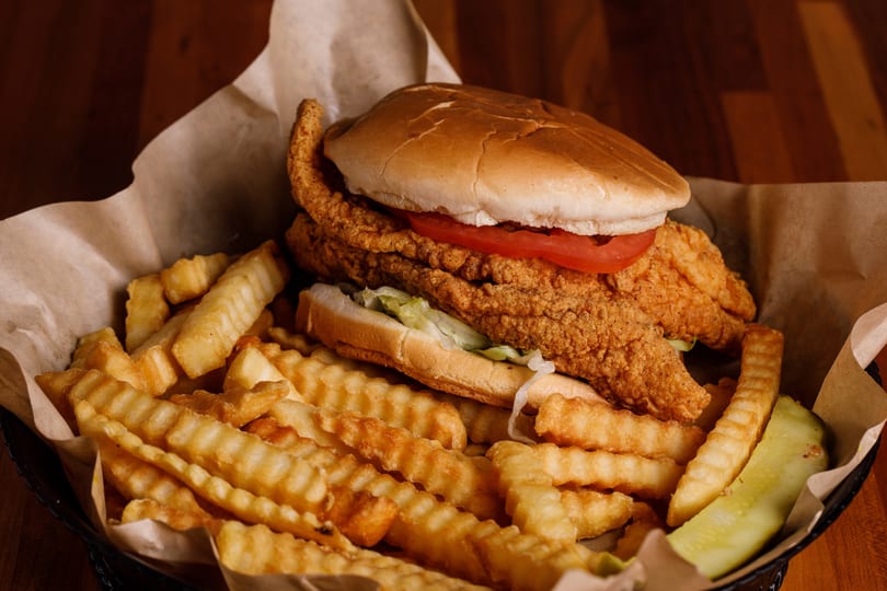 Crispy fried fish sandwich with lettuce and tomato, crinkle-cut fries, and a pickle spear.