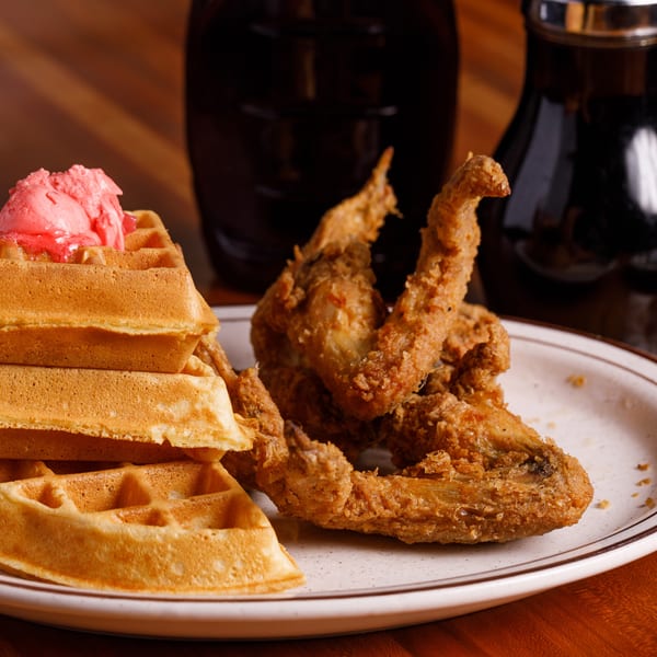 Crispy fried chicken wings and a stack of waffles with pink butter, with syrup in the background.