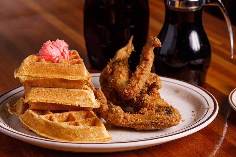 Crispy fried chicken wings and a stack of waffles with pink butter, with syrup in the background.
