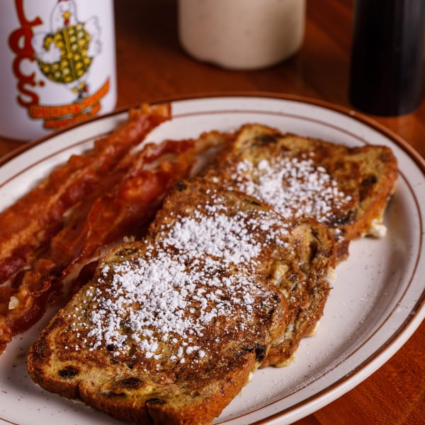 French toast with powdered sugar and crispy bacon on a white plate with a coffee mug.