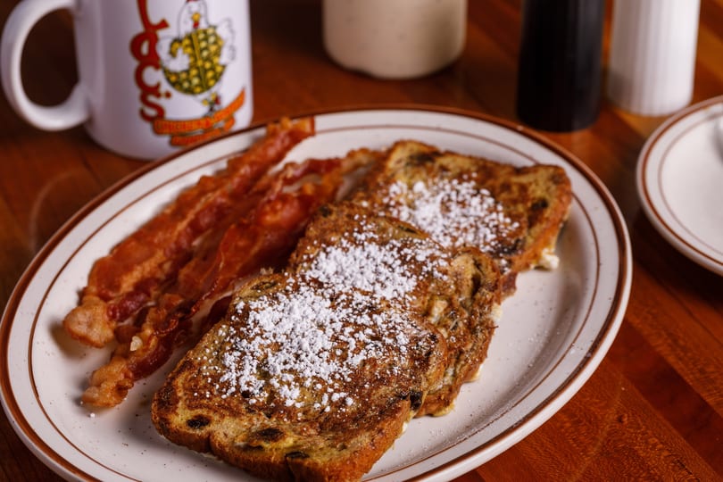 French toast with powdered sugar and crispy bacon on a white plate with a coffee mug.