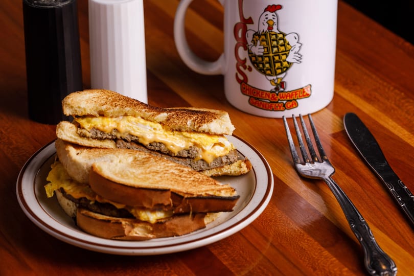 Breakfast sandwich with egg, cheese, and sausage on toast, with a branded mug and cutlery.