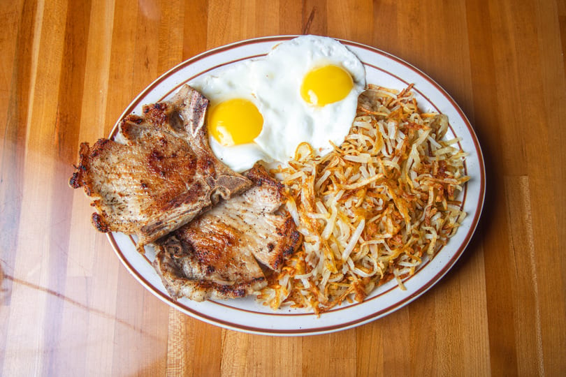 Two grilled pork chops, two fried eggs, and a pile of hash browns on a white plate.