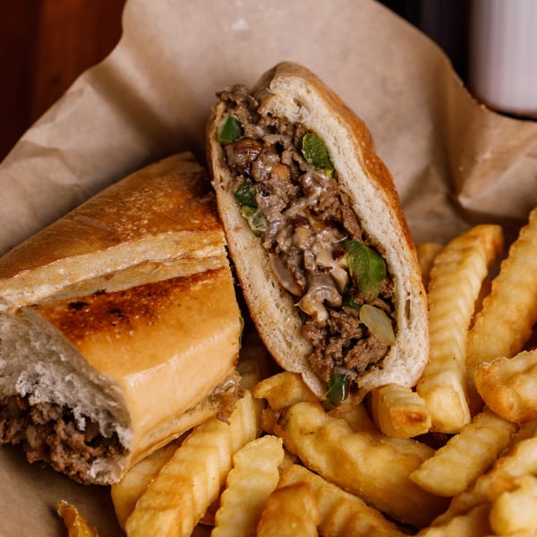 Cheesesteak sandwich, cut in half, with steak, peppers, onions, and mushrooms, served with crinkle-cut fries.