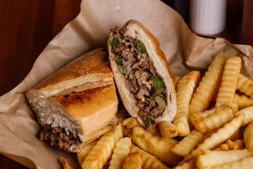 Cheesesteak sandwich, cut in half, with steak, peppers, onions, and mushrooms, served with crinkle-cut fries.