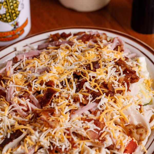 A large salad topped with shredded cheese, bacon bits, and ham, next to a white mug.