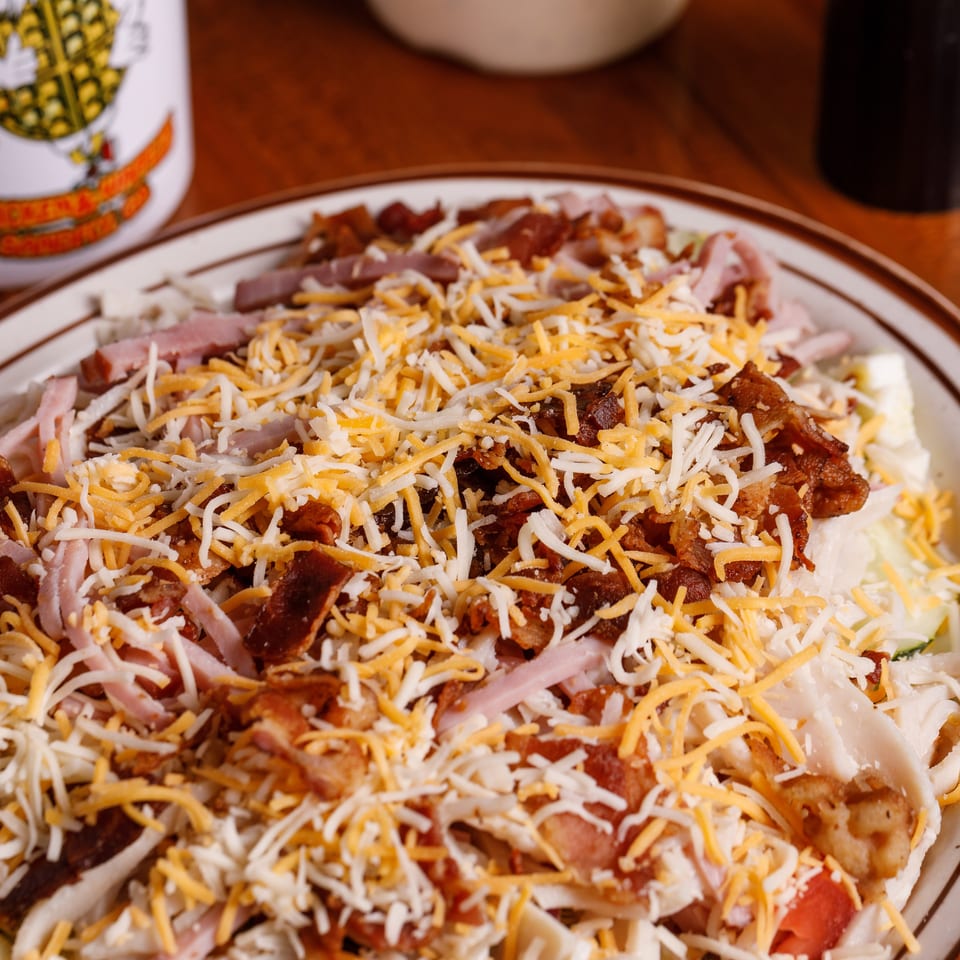 A large salad topped with shredded cheese, bacon bits, and ham, next to a white mug.