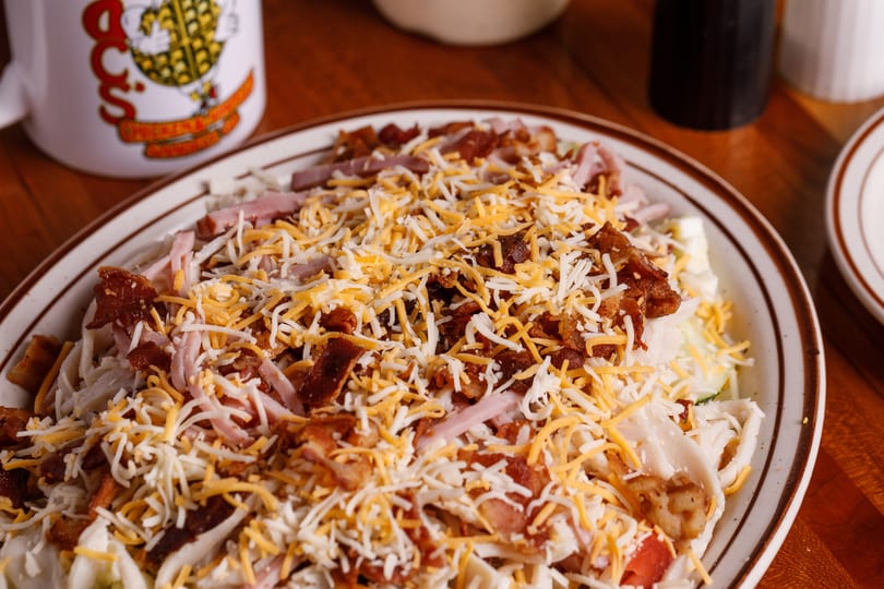 A large salad topped with shredded cheese, bacon bits, and ham, next to a white mug.