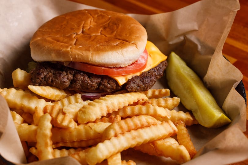 Cheeseburger with tomato, onion, crinkle-cut fries, and a pickle spear in a paper-lined basket.