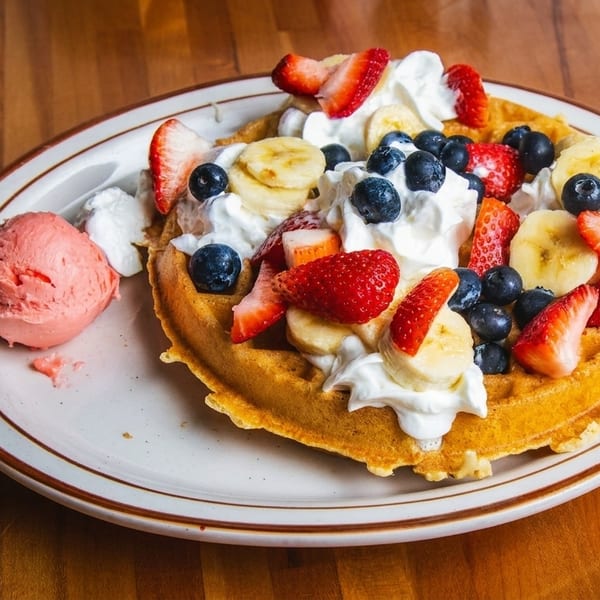 Golden waffle topped with whipped cream, strawberries, blueberries, bananas, and a scoop of pink ice cream.