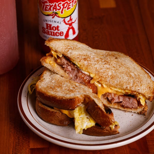 A grilled sandwich with sausage, egg, and cheese, served with Texas Pete hot sauce and a drink.