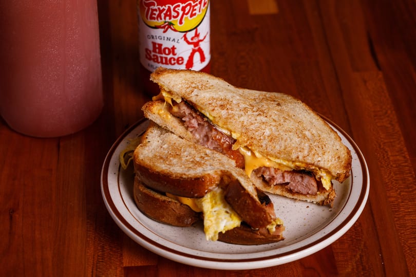 A grilled sandwich with sausage, egg, and cheese, served with Texas Pete hot sauce and a drink.